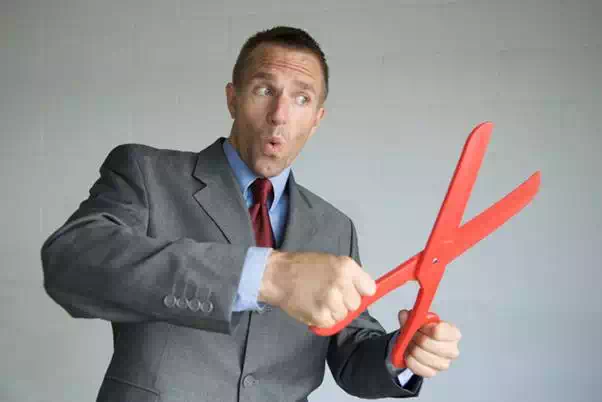 Businessman carefully trimming the budget with a big fat pair of red scissors against a light gray background