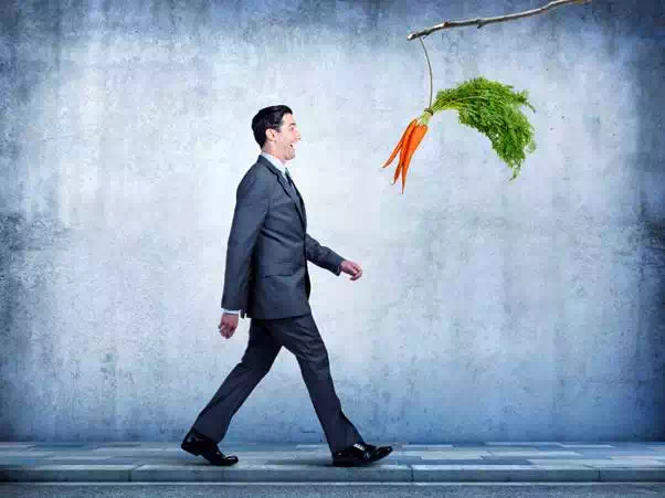 A businessman, walking along a sidewalk, happily follows a bunch of carrots that are dangling from a stick. The carrots are his motivation as he works his way through his day.